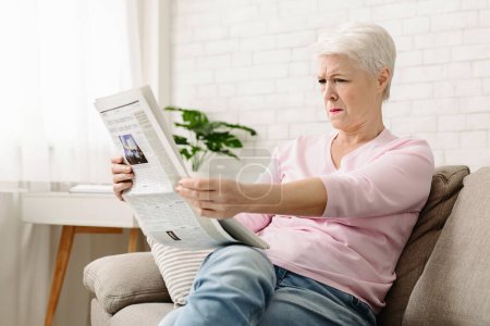 Une femme âgée est assise sur un canapé, absorbée par la lecture d'un journal. Elle se concentre sur le document, avec une expression sérieuse, ont des problèmes de vision. La chambre est doucement éclairée, et elle semble confortable.