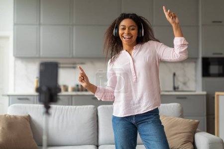 Eine energische Afroamerikanerin tanzt fröhlich, während sie mit ihrem Handy in ihrem modernen Zuhause live streamt