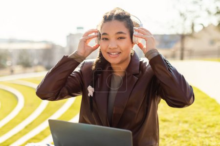 Téléchargez les photos : Une jeune étudiante joyeuse se tient dehors par une journée ensoleillée, ajustant son casque avec un doux sourire tout en tenant un ordinateur portable, peut-être en prenant une pause de l'étude - en image libre de droit