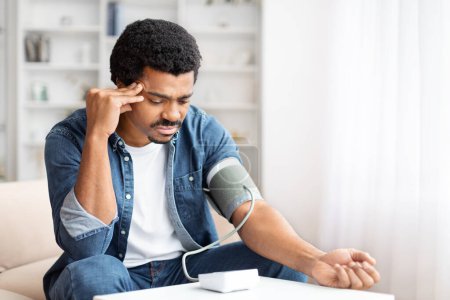 Ein erwachsener schwarzer Mann sieht besorgt aus, während er zu Hause ein digitales Blutdruckmessgerät benutzt, was auf eine Gesundheitsüberwachung oder mögliche Probleme hindeutet.