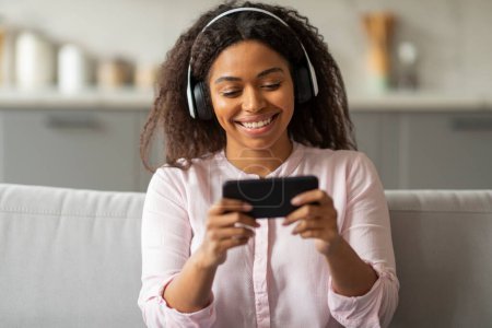 In a relaxed living room scene, a young black woman sits on a couch, deeply engrossed in her phone and wearing headphones, suggesting a moment of leisure and personal enjoyment at home