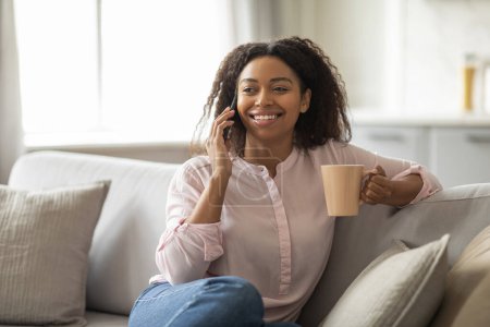 Téléchargez les photos : Joyeux jeune femme afro-américaine appréciant un appel téléphonique tout en tenant une tasse de café à la maison, assis sur le canapé, espace de copie - en image libre de droit