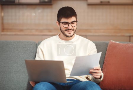 Ein Mann sitzt auf einem Sofa mit Notizbuch auf dem Schoß, konzentriert auf ein Blatt Papier in der Hand, Unternehmer, der von zu Hause aus arbeitet