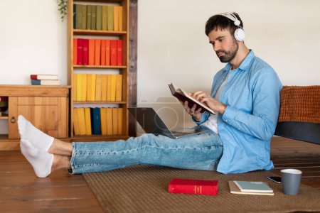 A focused young man reclines on the floor with his legs extended, engrossed in a book while wearing white headphones. His casual attire suggests comfort within a cozy home environment