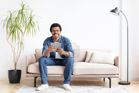 Joyeux homme afro-américain à la maison assis sur le canapé, en utilisant une tablette numérique, exprimant la communication et le bonheur
