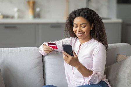 Téléchargez les photos : Femme afro-américaine assise sur le canapé, tenant une carte de crédit et un smartphone, représentant des transactions en ligne à la maison - en image libre de droit