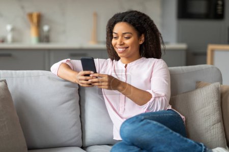 Téléchargez les photos : Une femme afro-américaine confortablement assise sur un canapé gris à la maison, s'engageant avec son smartphone, représentant une connexion avec les gens - en image libre de droit