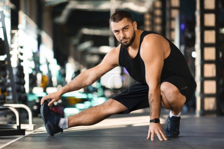Un homme est accroupi sur le sol dans une salle de gym. Il porte des vêtements d'entraînement et semble concentré sur sa routine d'entraînement. La salle de gym est bien équipée avec diverses machines d'exercice en arrière-plan.