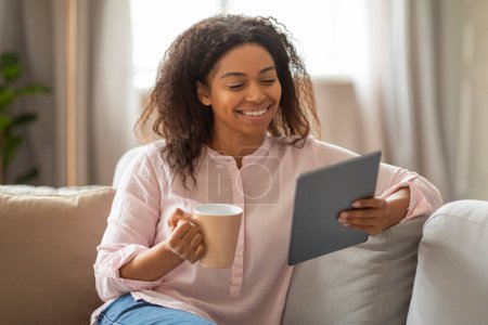Smiling African American woman enjoys a hot beverage while browsing a tablet at home, displaying relaxed digital interaction