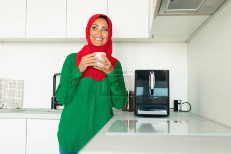 Muslimische Frau steht in einer Küche und hält eine Tasse Kaffee in der Hand. Die Küche ist gut beleuchtet, im Hintergrund sind verschiedene Utensilien und Geräte zu sehen..