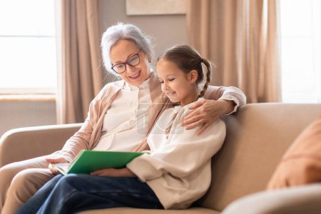 Una mujer mayor y una joven están sentadas en un sofá en casa, conversando mientras leen un libro juntas.