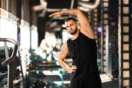 Un hombre está de pie en un gimnasio, sosteniendo su brazo en el aire. Aparece concentrado y decidido, mostrando fuerza y forma física.