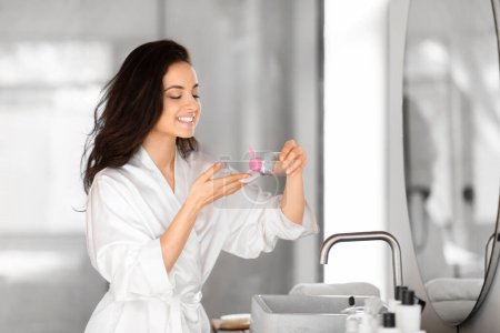 Téléchargez les photos : Une femme millénaire exubérante en peignoir blanc admire un produit de soins de la peau dans sa salle de bain bien équipée, le visage nettoyant - en image libre de droit