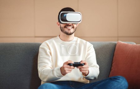 A young man sits on a cozy couch with a virtual reality headset on his head, holding a controller in his hands with a relaxed smile, seemingly enjoying a virtual reality game