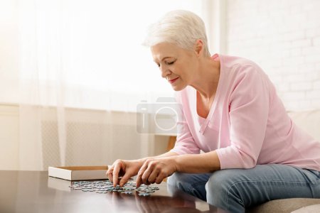 Senior woman is seated at a table, engrossed in playing with a puzzle. Concentration evident on her face as she pieces together the puzzle.