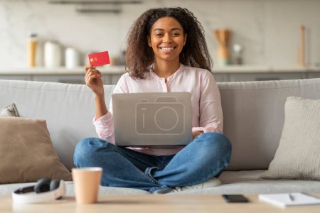Téléchargez les photos : Femme afro-américaine souriante tenant une carte de crédit tout en faisant des achats en ligne avec un ordinateur portable dans un cadre confortable à la maison - en image libre de droit