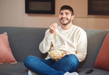 Photo for A man is seated comfortably on a couch, munching on a bowl of popcorn. He seems relaxed while enjoying his snack in the cozy setting. - Royalty Free Image