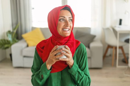 A cheerful woman wearing a red hijab and green blouse stands in a sunlit living room, holding a gray mug with both hands. She gazes out of the frame with a joyful expression