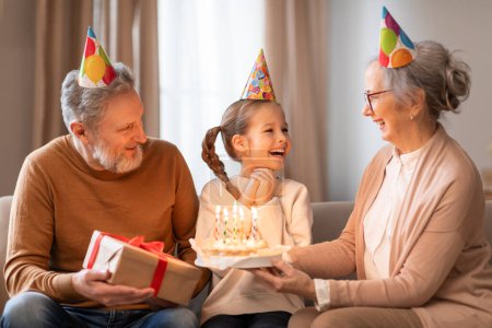 Photo for A young girl, wearing a festive party hat, gleefully receives a birthday cake from her grandmother, while an equally joyous grandfather looks on holding a gift - Royalty Free Image