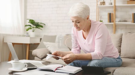 A senior woman with short white hair is seated at a cozy living space, meticulously examining financial papers. She appears concentrated, with a calculator, notebook, and laptop on the table