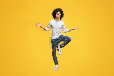 Funny young man sitting in mid air, balancing on one leg, orange studio background