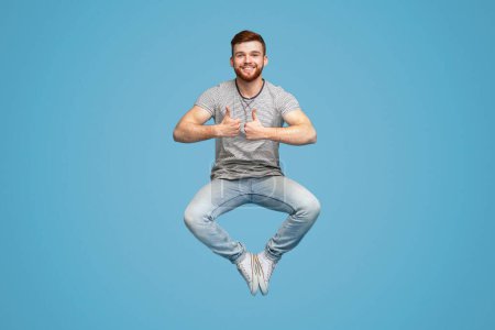 Enjoy life moments. Joyful redhaired man jumping in air, demonstrating thumbs up gesture, blue studio background, full length, free space