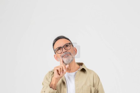 A senior man with a beard and glasses is standing confidently in front of the camera, touching his face and looking above at copy space, white background