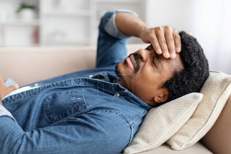 Captured in a serene home environment, this image portrays a young african american guy lying on couch at home, touching head, suffering from migraine