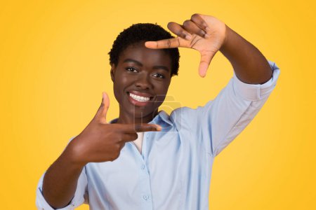 Foto de Una alegre joven afroamericana está haciendo un marco con sus manos, simulando un visor de cámara, sobre un cálido fondo amarillo - Imagen libre de derechos