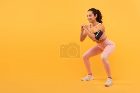 Eine junge Frau mit nach hinten gebundenen Haaren macht ein Workout, demonstriert eine gehockte Position, trägt Trainingsbekleidung, darunter einen hellrosa Sport-BH und Leggings mit weißen Turnschuhen, Kopierraum