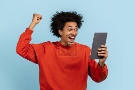 Ein ekstatischer afrikanisch-amerikanischer Typ mit einem digitalen Tablet feiert, isoliert auf blauem Grund, ideal, um Erfolg und Leistung auszudrücken