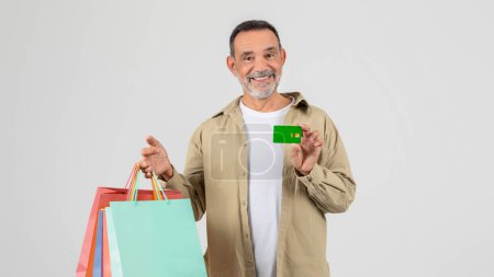 Foto de Un hombre mayor parado sosteniendo bolsas de compras en una mano y una tarjeta de crédito en la otra. Parece estar en una zona comercial, posiblemente un centro comercial, listo para hacer compras.. - Imagen libre de derechos