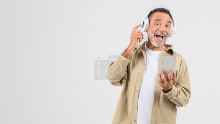 Foto de Un hombre mayor sostiene un teléfono celular y usa auriculares, escucha música, disfruta de canciones favoritas sobre fondo blanco, copia espacio - Imagen libre de derechos