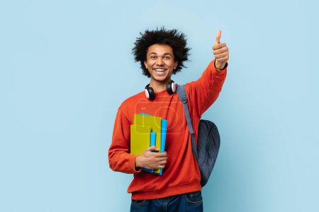 Téléchargez les photos : Un homme afro-américain joyeux avec écouteurs, tenant des livres et montrant un pouce vers le haut symbolise le succès de l'éducation et la positivité parmi les étudiants zoomer - en image libre de droit