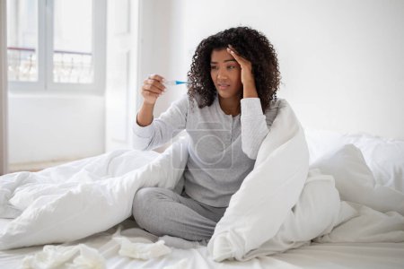 Eine junge hispanische Frau in eine weiße Decke gehüllt sitzt mit besorgtem Gesichtsausdruck im Bett, während sie auf ein Thermometer blickt..