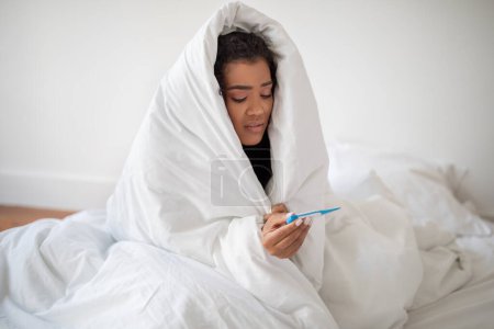 Photo for A young Hispanic woman, wrapped cozily in a white blanket, is sitting on her bed while intently looking at a thermometer in her hand, possibly checking for a fever - Royalty Free Image