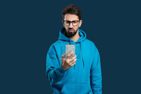 Photo for A young, bearded man wearing glasses and a blue hoodie stands against a solid blue background, looking at his smartphone with a skeptical and slightly confused expression. - Royalty Free Image