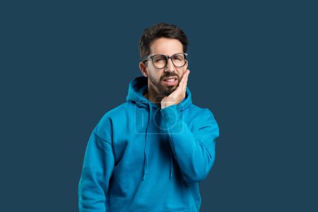 A man wearing glasses and a blue hoodie is standing against a neutral blue backdrop, holding his cheek and jaw with an expression of discomfort indicative of dental pain or a toothache.