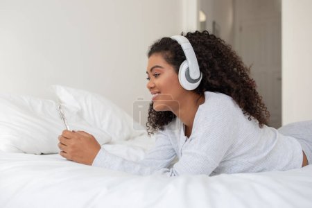 Photo for A young Hispanic woman with curly hair lies comfortably on a white bed, dressed in a casual white outfit, enjoying music through her white headphones, with a smartphone in her hand. - Royalty Free Image