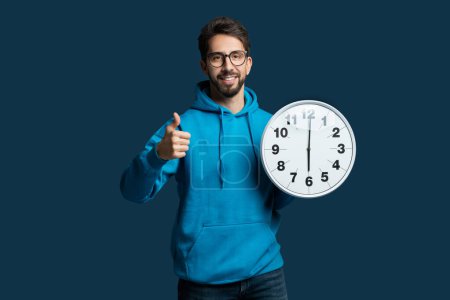 Un homme portant un sweat à capuche bleu est représenté tenant une horloge dans ses mains, regardant l'heure. Le visage des hommes est sans expression, et l'accent est mis sur l'horloge qu'il tient.