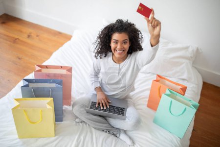 A Hispanic woman is seated on a bed, holding a credit card in one hand and a laptop in the other, possibly making an online purchase or managing finances, top view