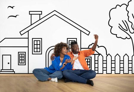 Foto de Amantes marido de familia negro y la esposa sentados sobre dibujado en la casa de pared blanca, sosteniendo la llave y sonriendo. Bienes raíces, concepto de hipoteca - Imagen libre de derechos