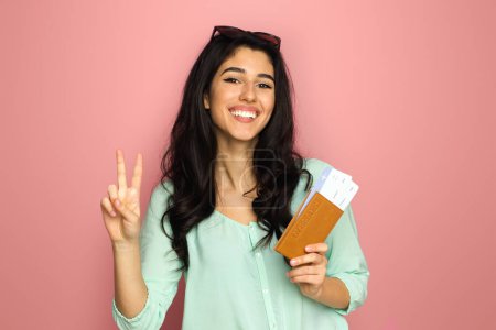 Eine fröhliche junge Frau aus dem Mittleren Osten, bereit für die Reise, steht vor rosa Hintergrund, hält ihren Pass und ihre Bordkarte in der Hand und blinkt mit einem strahlenden Lächeln auf ein Friedenszeichen..