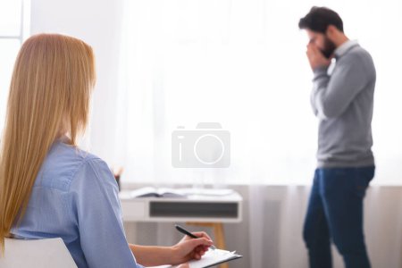 Téléchargez les photos : A man stands with his hand on his forehead in a posture of distress or deep thought while facing away from woman therapist - en image libre de droit