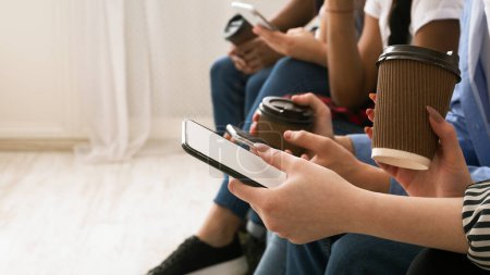 Un grupo de adolescentes están sentados juntos, con su enfoque en sus teléfonos inteligentes portátiles. Un individuo sostiene una taza de café de papel, lo que sugiere un ambiente social relajado, recortado
