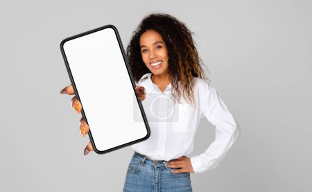 A cheerful young black woman with curly hair is holding a smartphone with a blank screen towards the camera.
