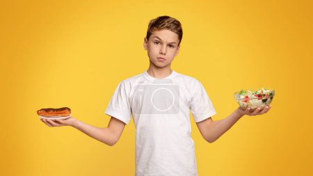 Foto de Problema de nutrición correcta y chatarra. Triste chico adolescente elegir entre ensalada de verduras y chocolate eclair, fondo de estudio naranja - Imagen libre de derechos