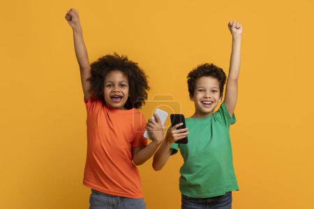 Photo for Two cheerful African American young kids are seen against a vibrant yellow backdrop, with arms triumphantly raised in the air, holding cell phones - Royalty Free Image
