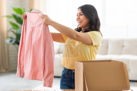 Eine fröhliche junge Frau aus dem Nahen Osten steht in einem sonnenbeschienenen Wohnzimmer und hält ein rosafarbenes Hemd mit zufriedenem Gesichtsausdruck in der Hand, während sie es aus einem Karton auspackt.