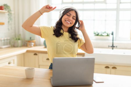 Photo for A cheerful young middle eastern woman is seen with closed eyes and headphones on, dancing slightly with her arms raised in joy as she listens to music on laptop, kitchen interior - Royalty Free Image